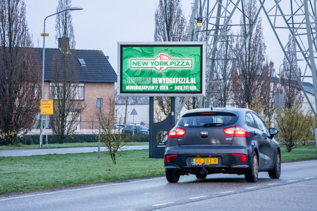 Een auto rijdt langs een Q-LED TV op de middenberm, met reclame van een pizzaverkoper.